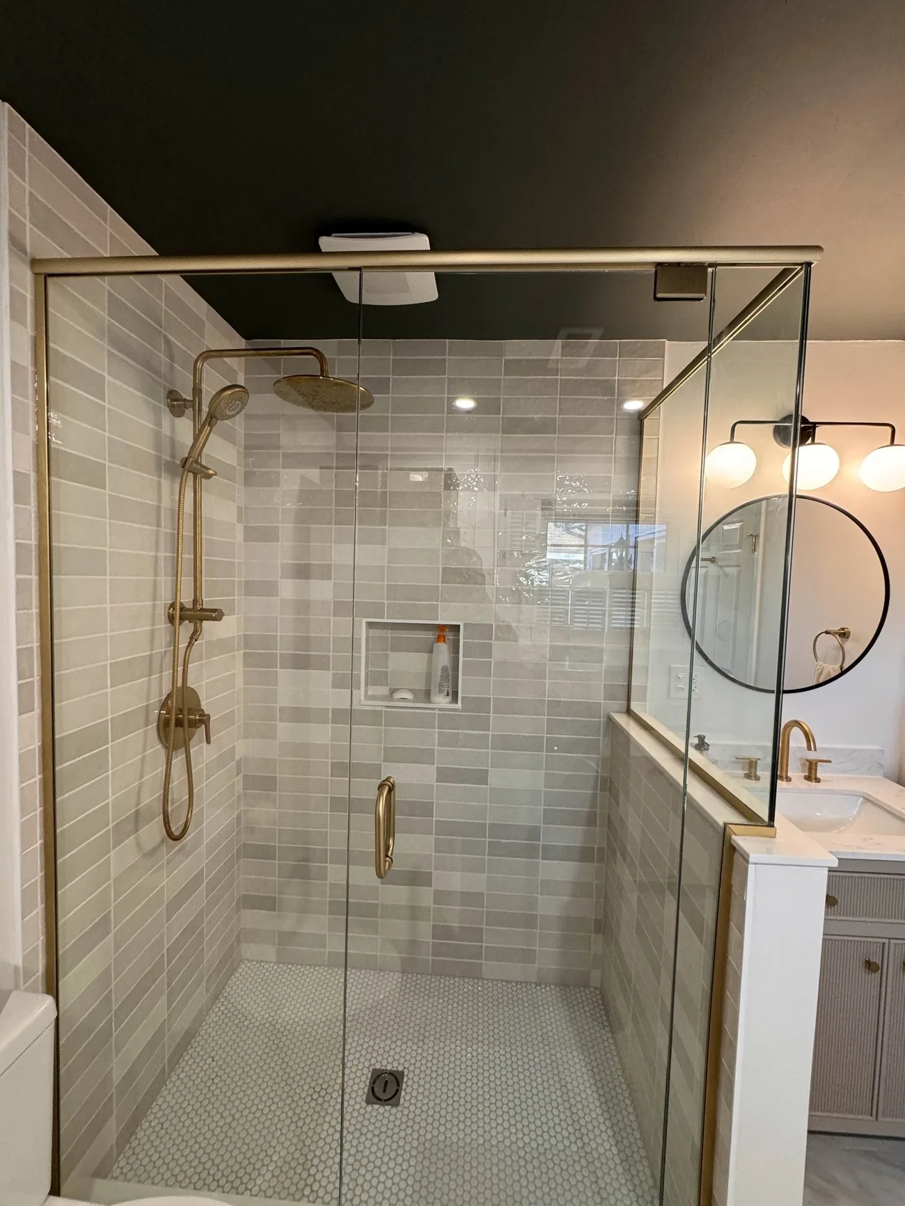 A finished walk-in shower with brass fixtures, glass panels and a round mirror in a remodeled bathroom.